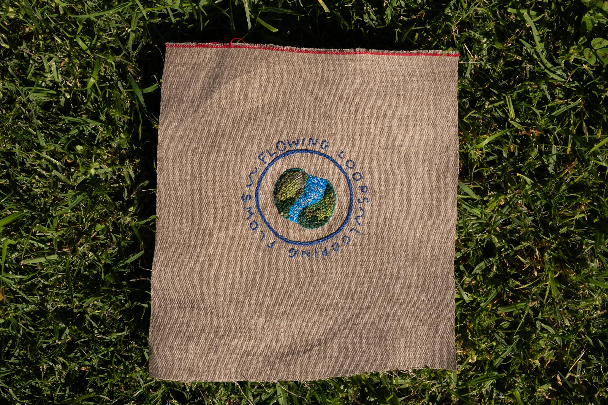 The Flowing Loops Logo is a photo of hand embroidered words "Flowing Loops Looping Flows" and blue depictions of a river flanked by greenery on rough brown canvas, with green grass as a background
